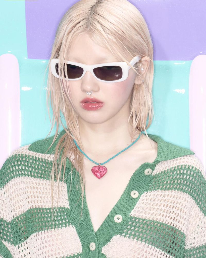 A young adult woman with platinum blonde hair wears sunglasses and a green striped cardigan in a studio portrait against a pastel background.