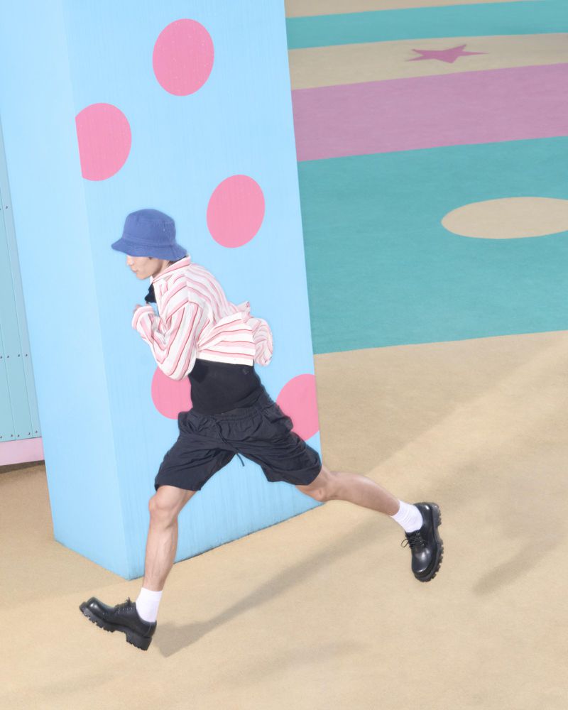 Person mid-stride in a light striped shirt, dark shorts, and blue bucket hat against a colorful polka dot and striped background.