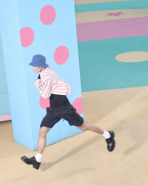 Person mid-stride in a light striped shirt, dark shorts, and blue bucket hat against a colorful polka dot and striped background.