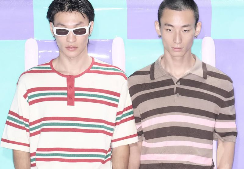Two adult men stand indoors against a striped, colourful backdrop, wearing casual shirts and stylish sunglasses. Fashion photography.