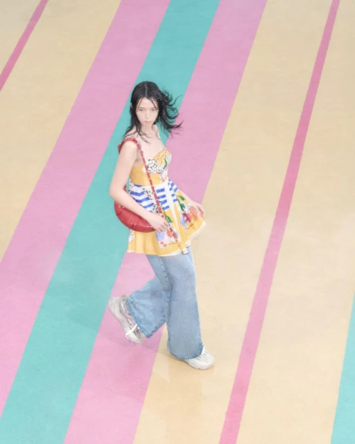 Woman in a patterned mini dress and blue jeans poses with a red purse on a striped pastel surface, creating a colorful fashion image.