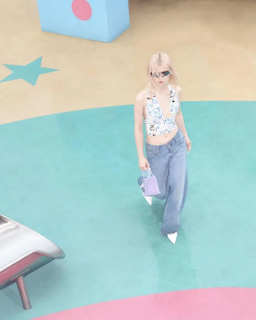 Fashionable blonde woman walks in an abstract colorful set, wearing a white top, baggy jeans, sunglasses, carrying a small purple purse.