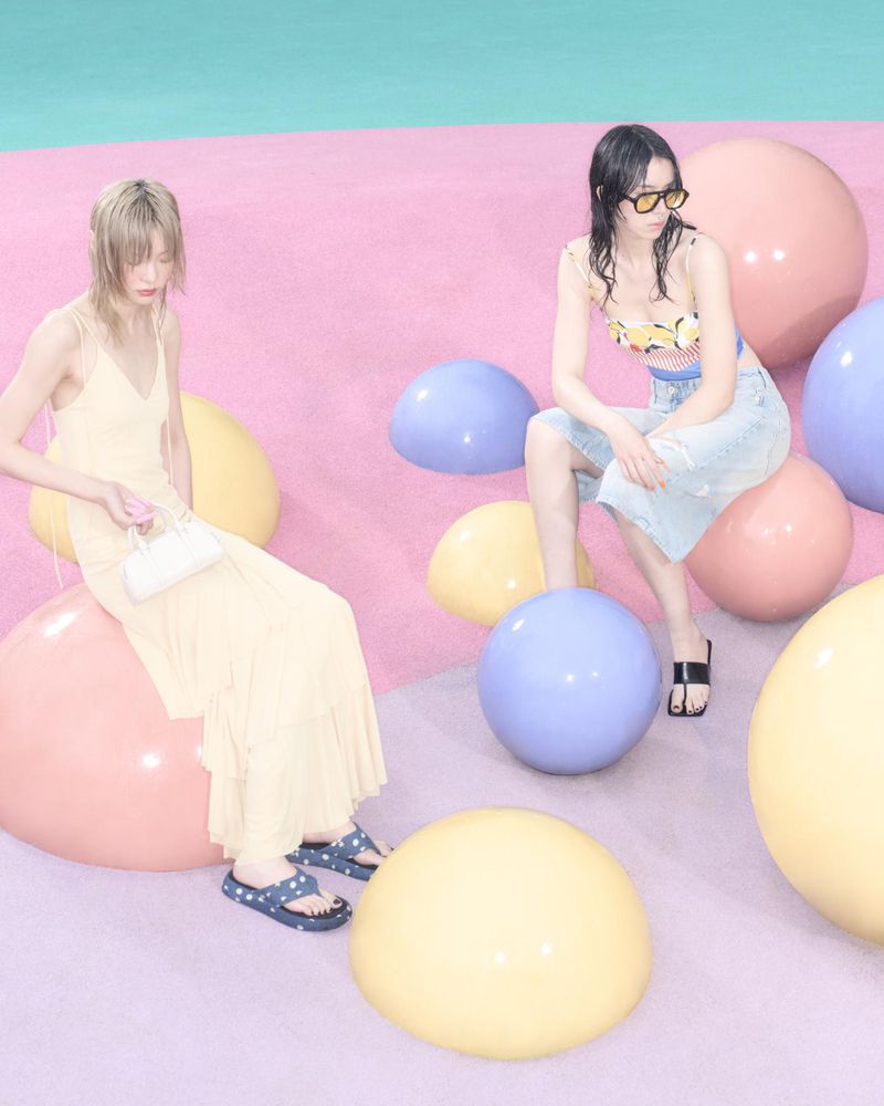 Two women pose while sitting on colorful spheres in a studio setting, showcasing fashion and style with soft lighting and pastel tones.