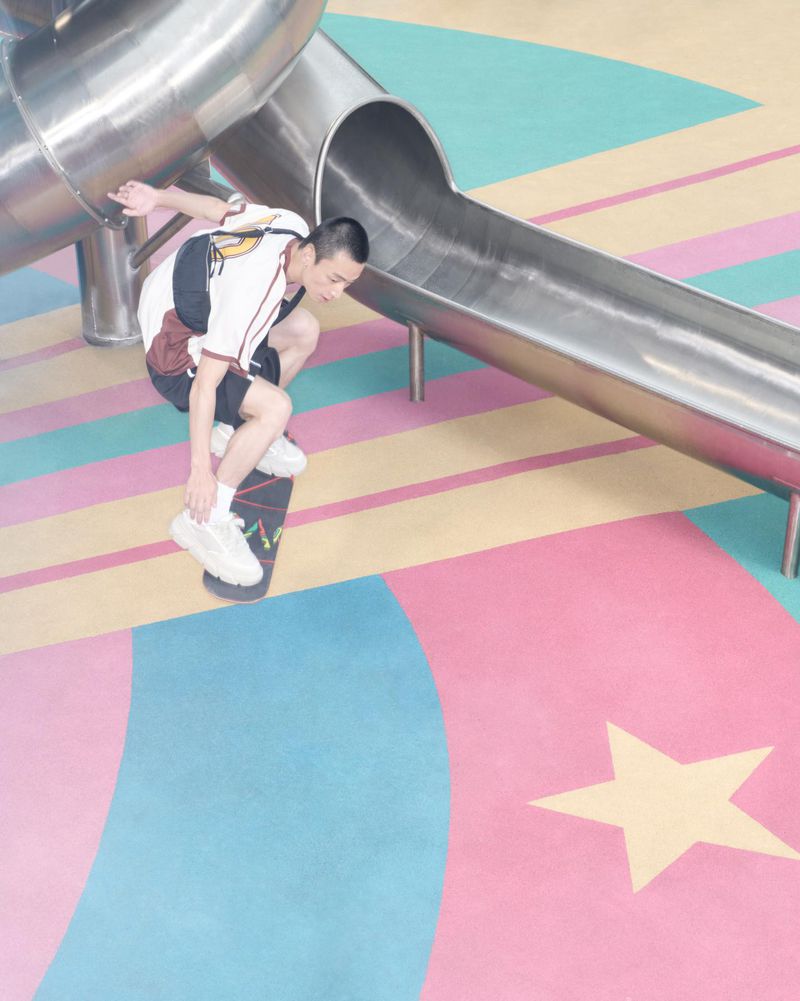 Young man performs skateboard trick near a playground slide wearing white sneakers, black shorts, and a matching top in an outdoor setting.