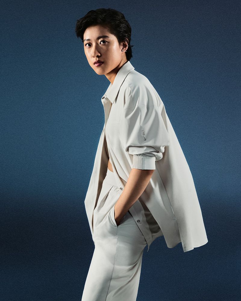 Portrait of a young adult woman in a light neutral-colored outfit with an unbuttoned shirt, with hands in pockets, looking at the camera.