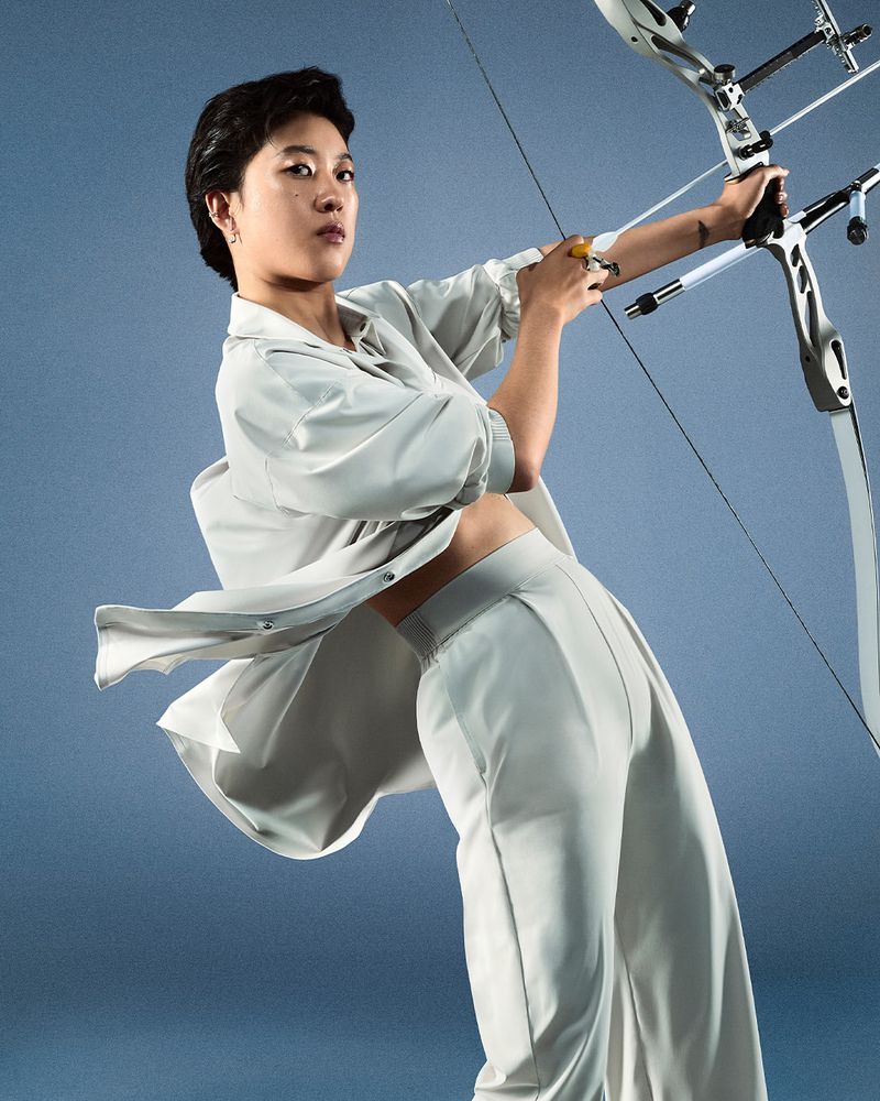 Athletic woman archer poses with modern bow and arrow in a studio setting. She wears a matching grey crop top and pants outfit.