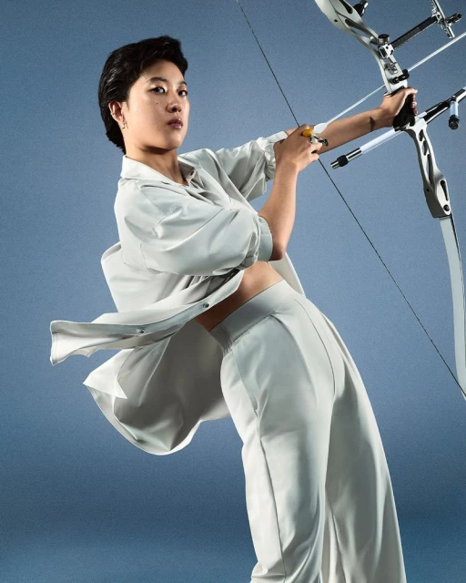 Athletic woman archer poses with modern bow and arrow in a studio setting. She wears a matching grey crop top and pants outfit.