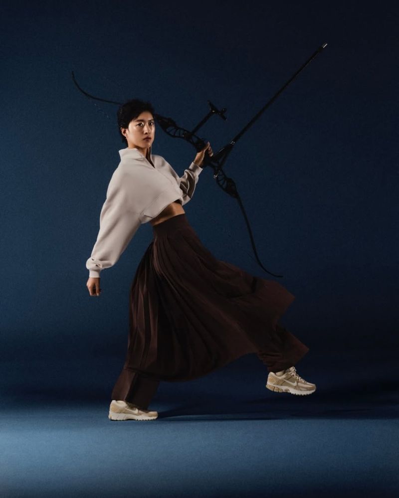 Young woman in athletic pose holding black recurve bow wearing streetwear clothing in a photo studio against a dark blue background.