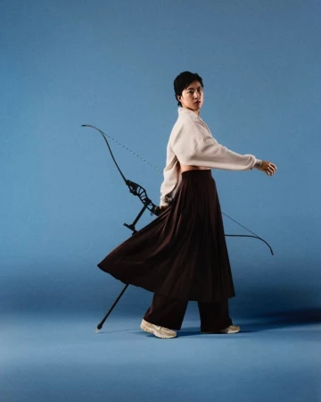 Woman with an archery bow poses in a studio setting, wearing a long skirt, cropped top, and sneakers against a solid blue backdrop.
