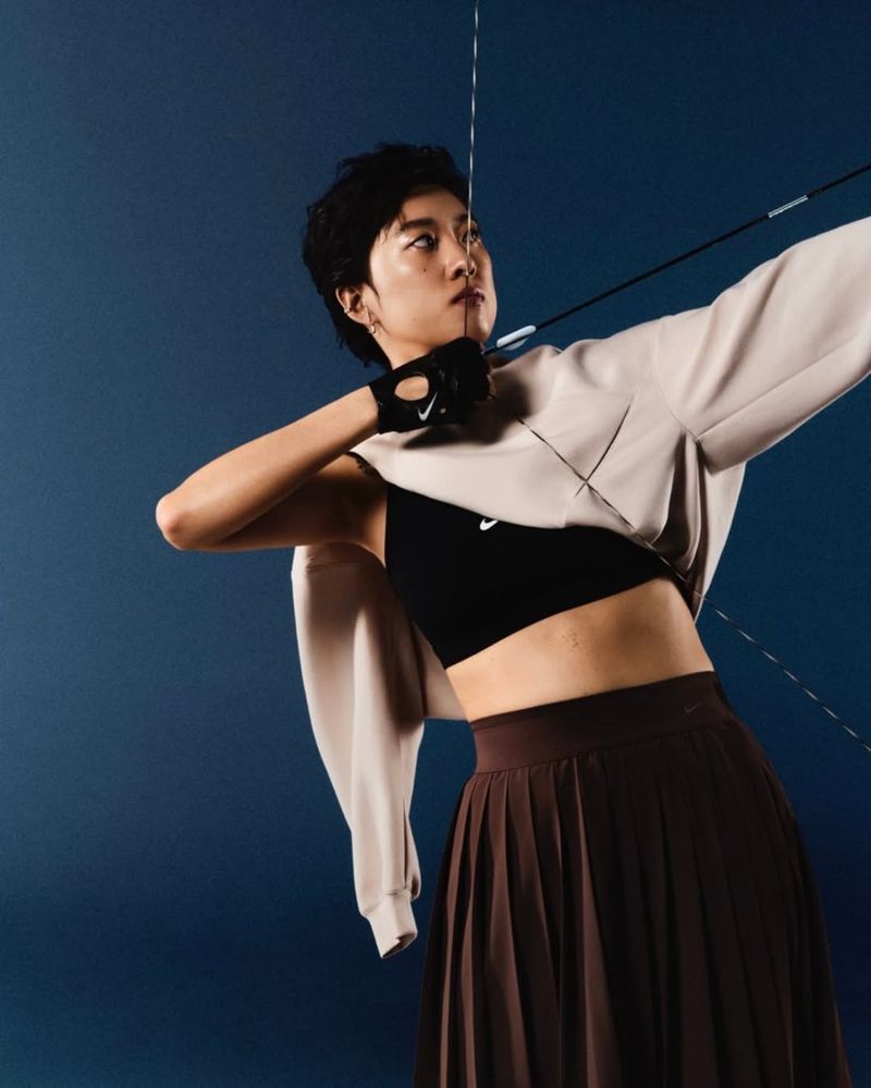 Athletic woman in sports bra and skirt aims an archery bow and arrow against a dark blue background in a studio shot.