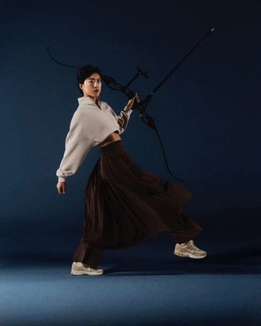 An San holding a recurve bow in a dynamic mid-stride pose, wearing a cropped sweatshirt and flowing brown trousers against a blue backdrop.