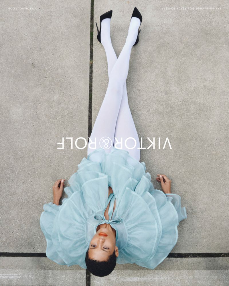 Model Laiza de Moura lying on concrete pavement wearing a tiered light blue ruffled dress, white tights, and black high heels.