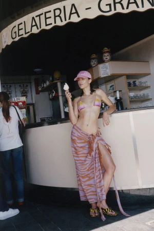 Model wearing a pink bikini top, patterned purple sarong, and pink cap holding an ice cream cone outside an Italian gelateria.