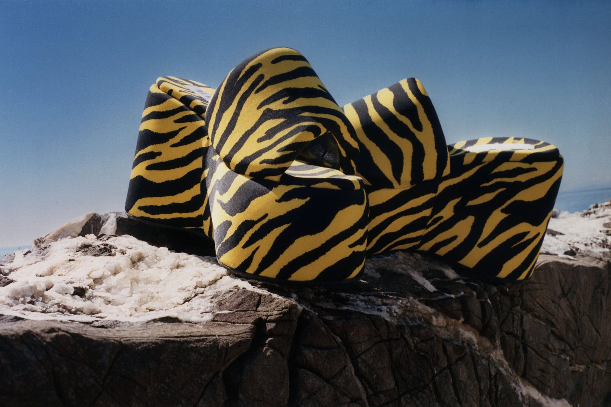 A pair of yellow and black tiger-print platform wedge sandals resting on rugged coastal rocks under a clear blue sky.
