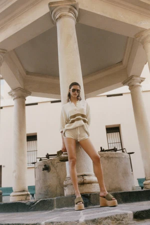 Model Adele Aldighieri wearing cream knit polo and shorts with platform sandals, leaning against a classical stone column.