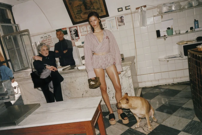 Model Adele Aldighieri wearing a pink striped wrap shirt and shorts set with platform sandals standing by a dog in a tiled shop interior.