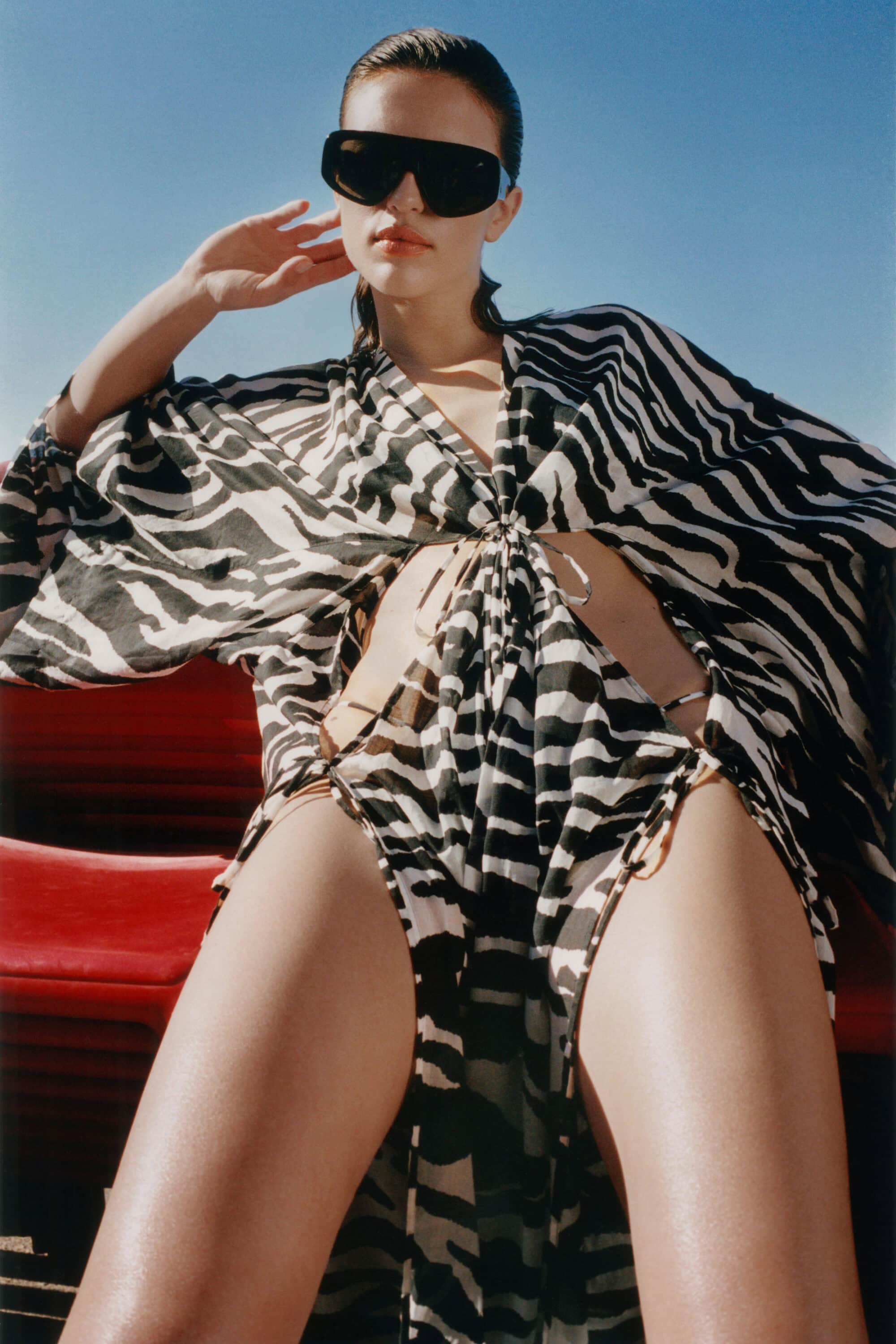 Low-angle shot of a woman wearing a zebra-print kaftan dress and oversized black sunglasses under a clear blue sky.