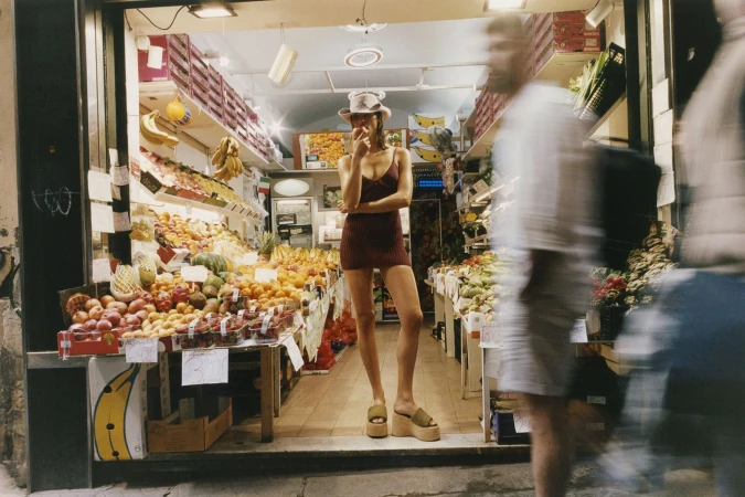 Model Adele Aldighieri eating fruit in a fitted brown mini dress and sun hat inside a brightly lit produce market.