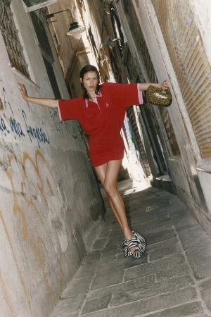 Model posing in a red polo mini dress and zebra-print platform heels with outstretched arms in a narrow stone alley.