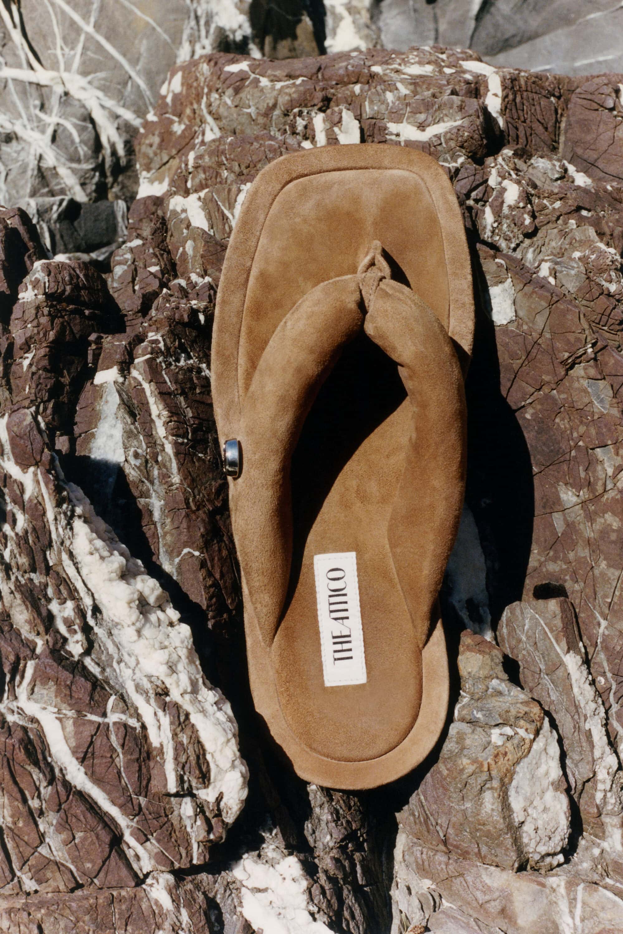 Brown suede padded thong sandal by The Attico resting on a textured, veined rock surface.