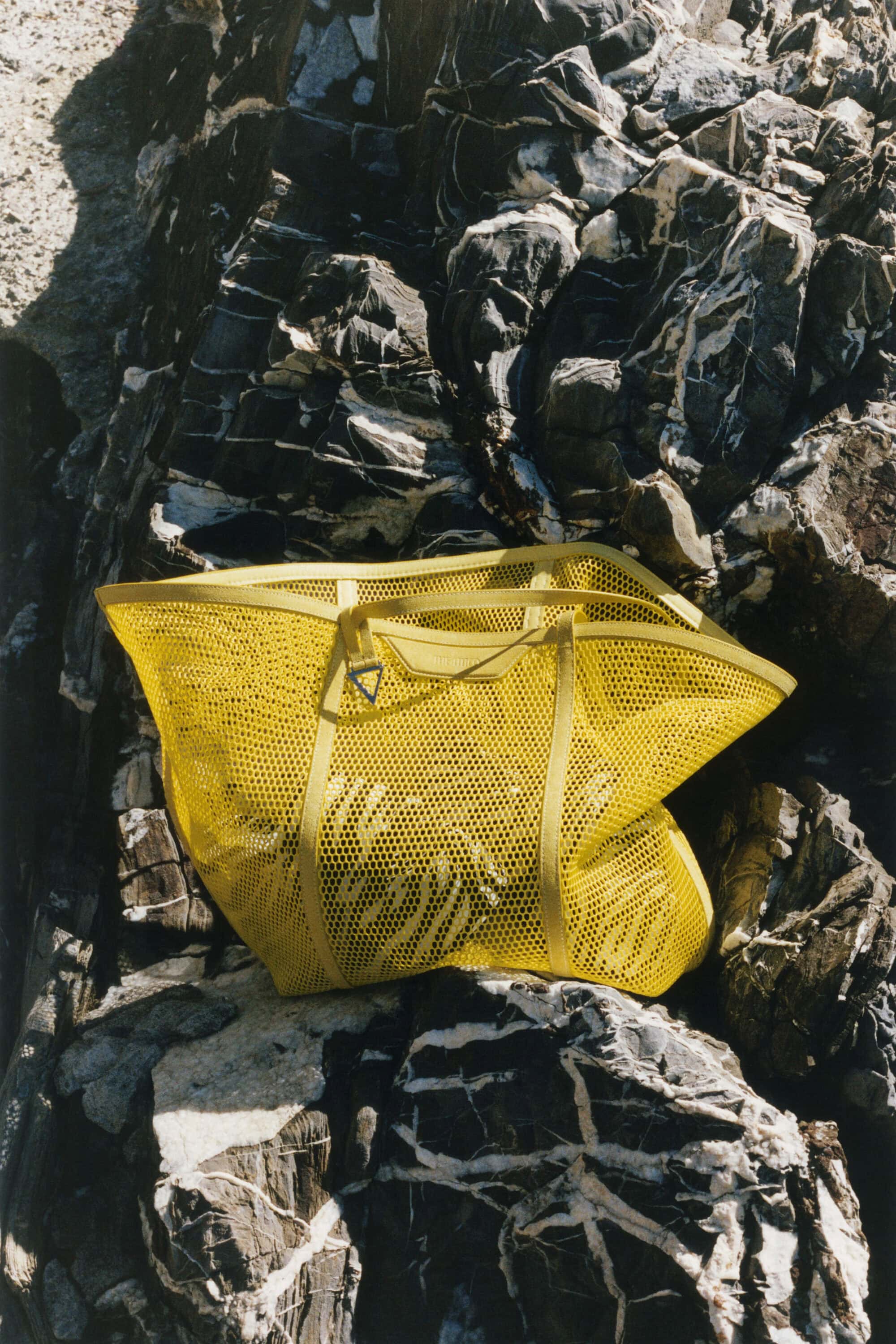 Bright yellow mesh tote bag resting on textured dark rocks with white mineral veins in harsh sunlight.