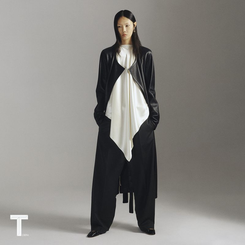 Model Sijia Kang wearing a draped white top, long black coat, and wide trousers against a grey studio backdrop.