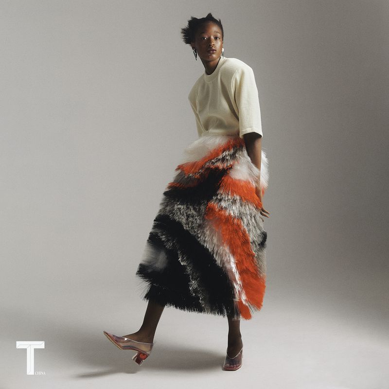Model posing in a cream short-sleeve top and tiered orange, black, and white fringe skirt against a grey studio backdrop.
