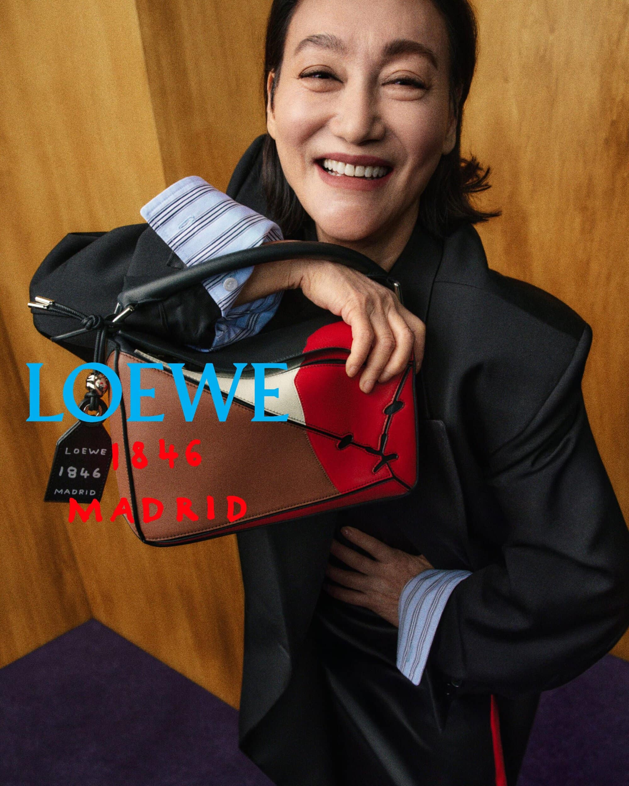 Kara Wai smiles while holding a multicolor Loewe leather handbag wearing an oversized black blazer against wood paneling.