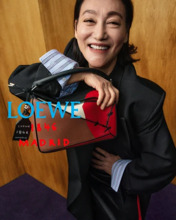 Kara Wai smiles while holding a multicolor Loewe leather handbag wearing an oversized black blazer against wood paneling.