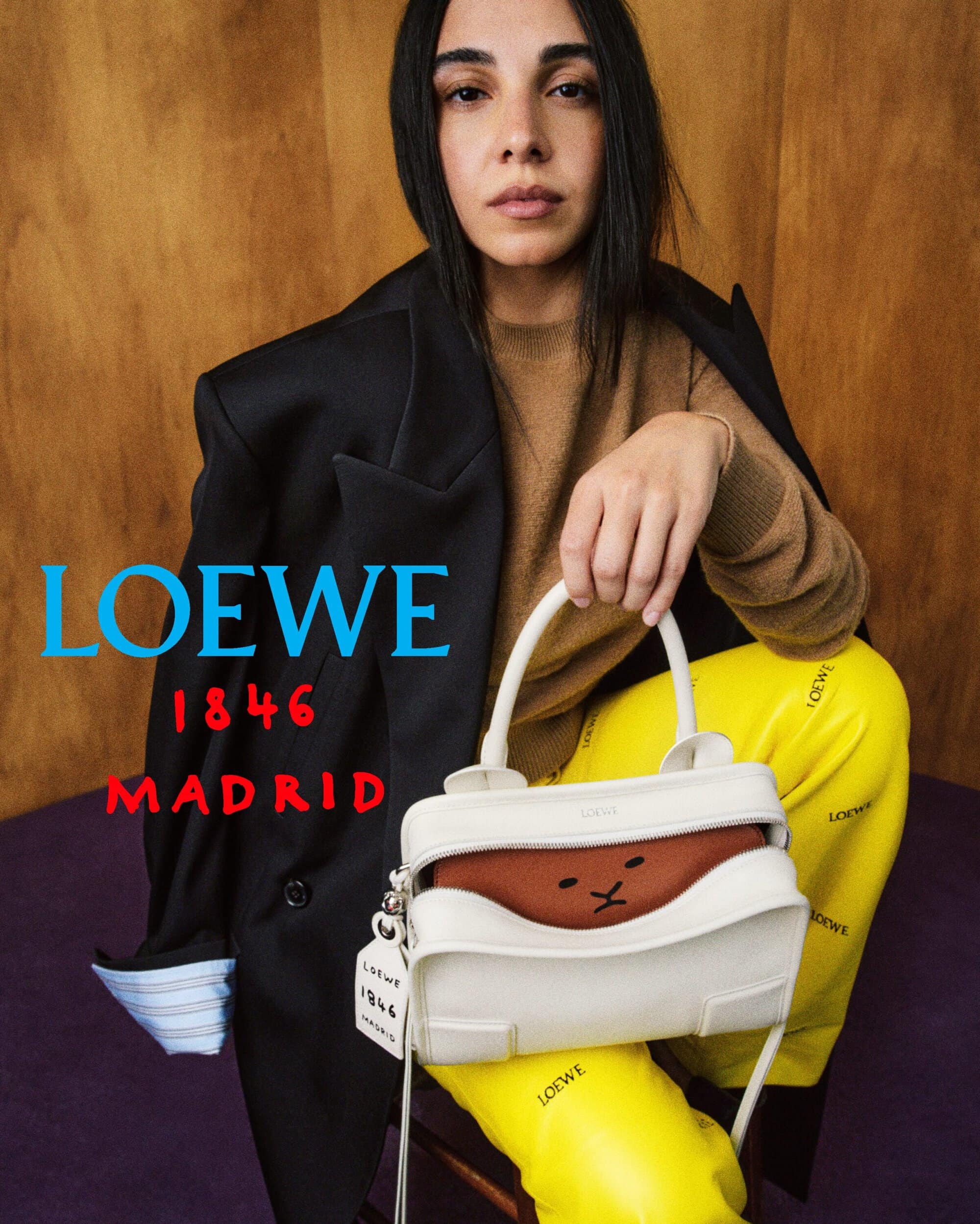 Salma Abu Deif posing with a white Loewe leather handbag while wearing yellow branded trousers and a draped black blazer.