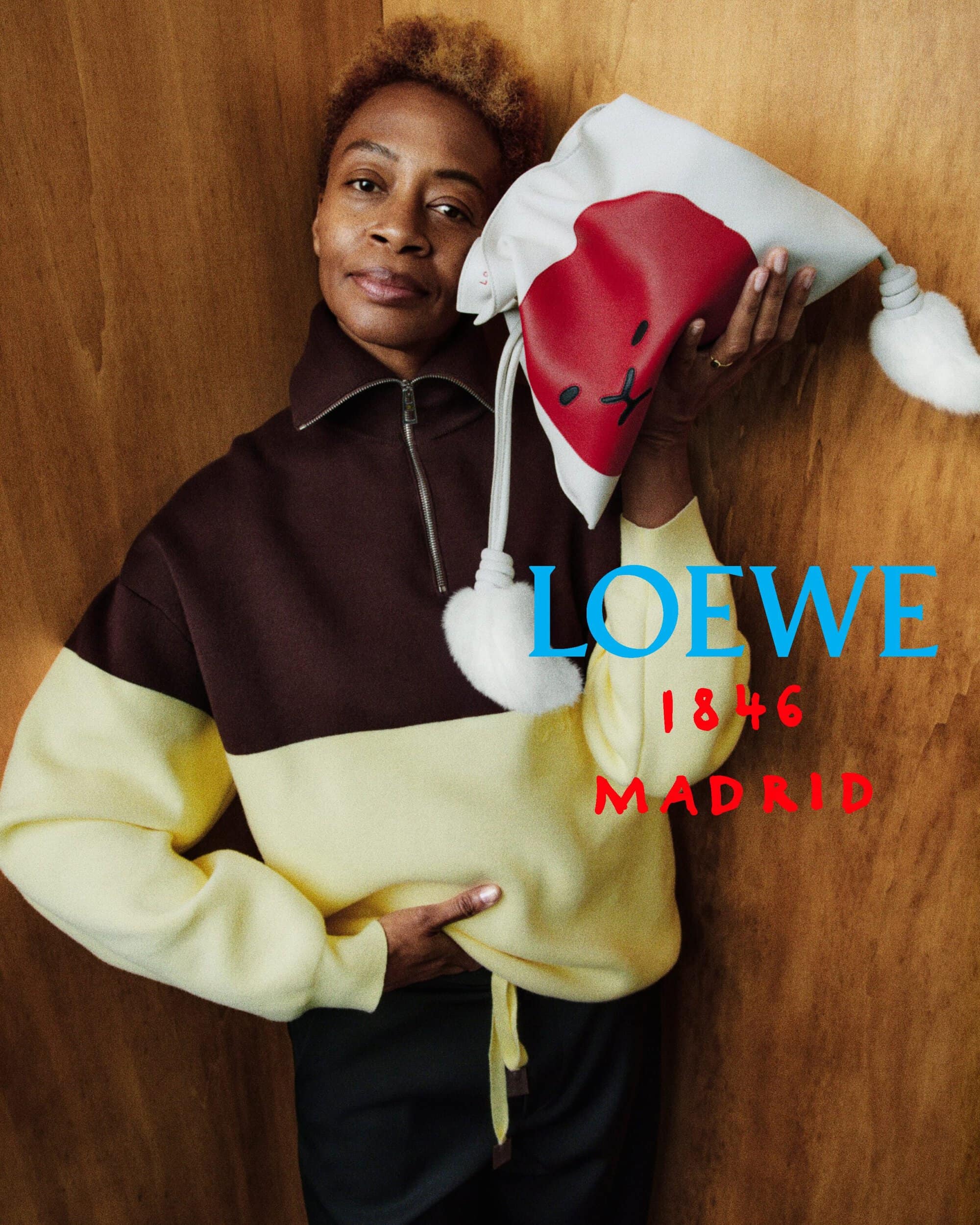 Kara Walker holding a white and red Loewe pouch with pom-poms while wearing a brown and yellow pullover against wood paneling.