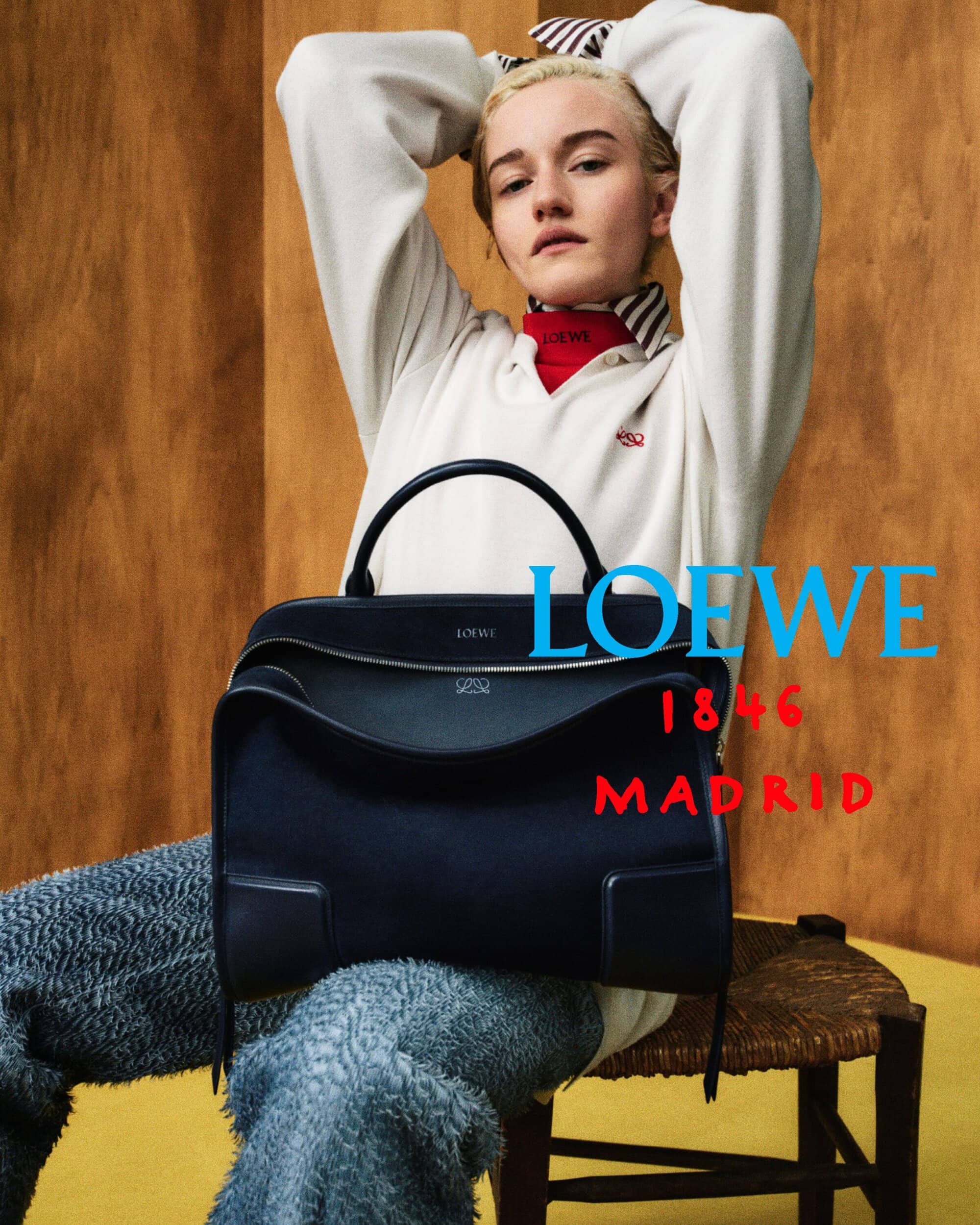 Model seated on a wooden chair holding a large dark Loewe handbag against a wood-panel wall with campaign typography.