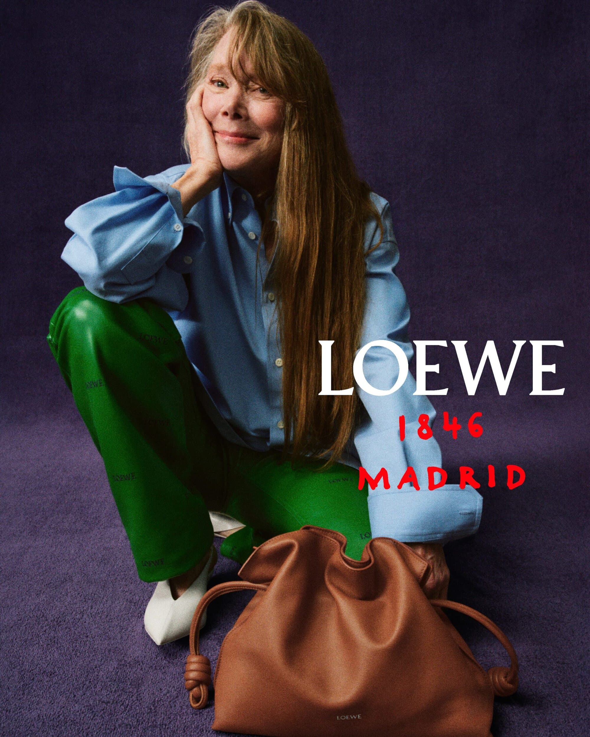 Sissy Spacek wearing a blue shirt and green pants squatting next to a brown leather handbag against a textured purple background