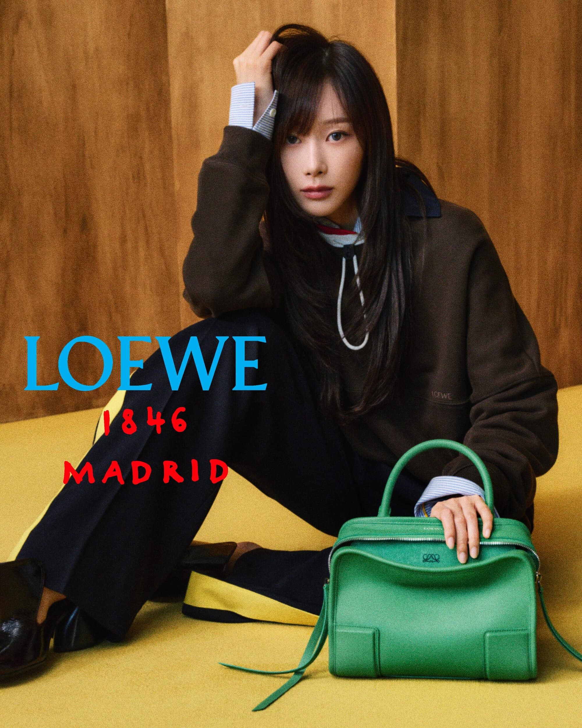 Giselle seated on a yellow floor in front of a wood wall, resting her hand on a vibrant green leather Loewe handbag.