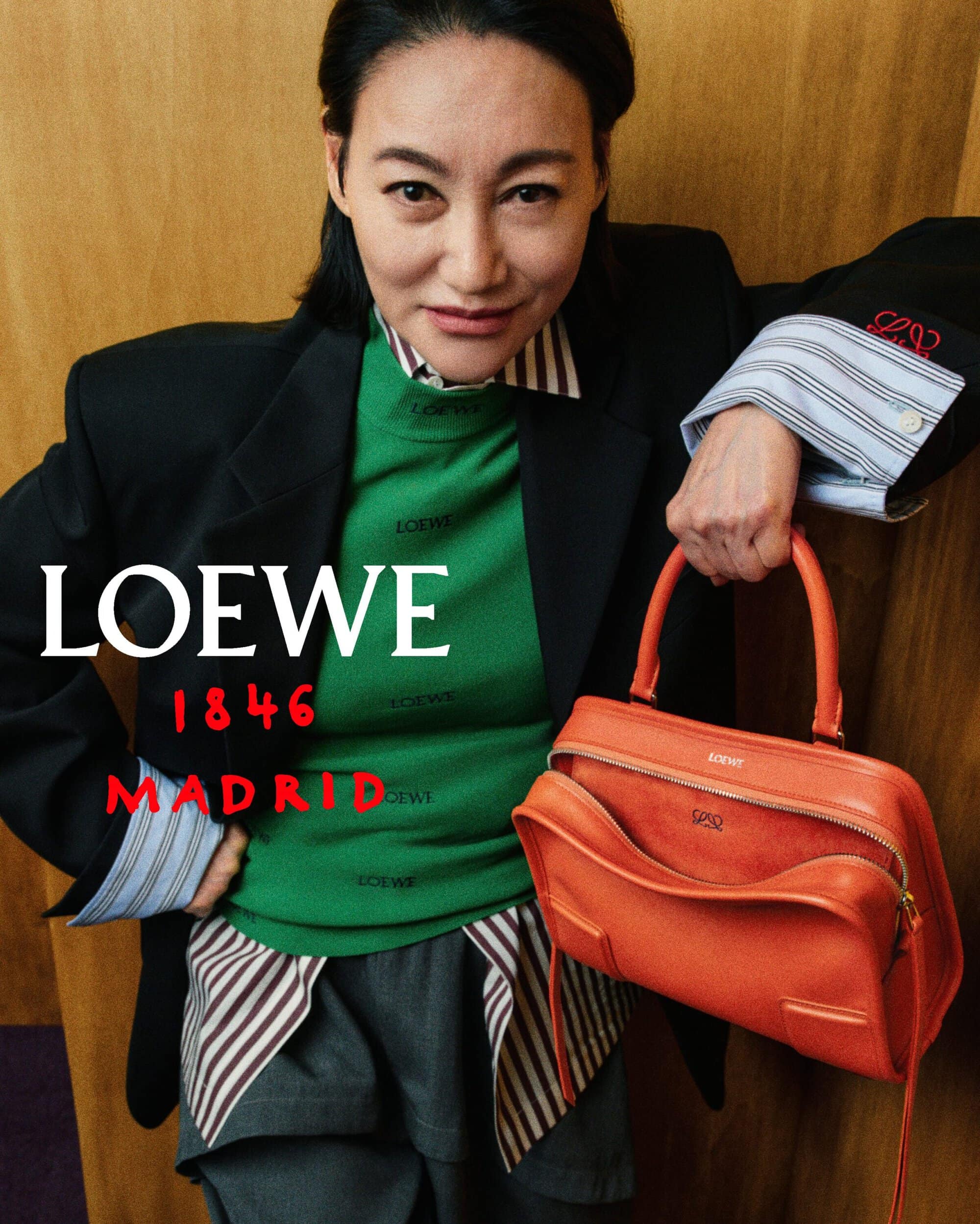 Kara Wai wearing a black blazer and green top while holding an orange Loewe leather handbag against a wood wall.