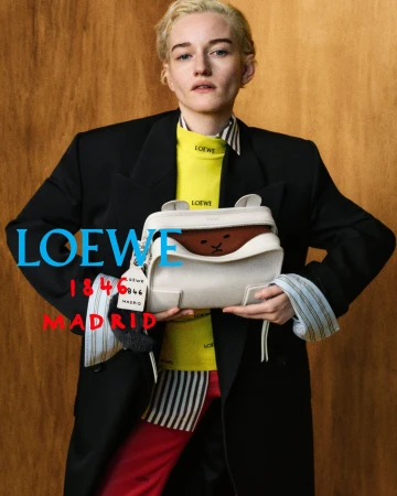 A model in an oversized black blazer and yellow top holds a white Loewe leather handbag against a wood panel backdrop.