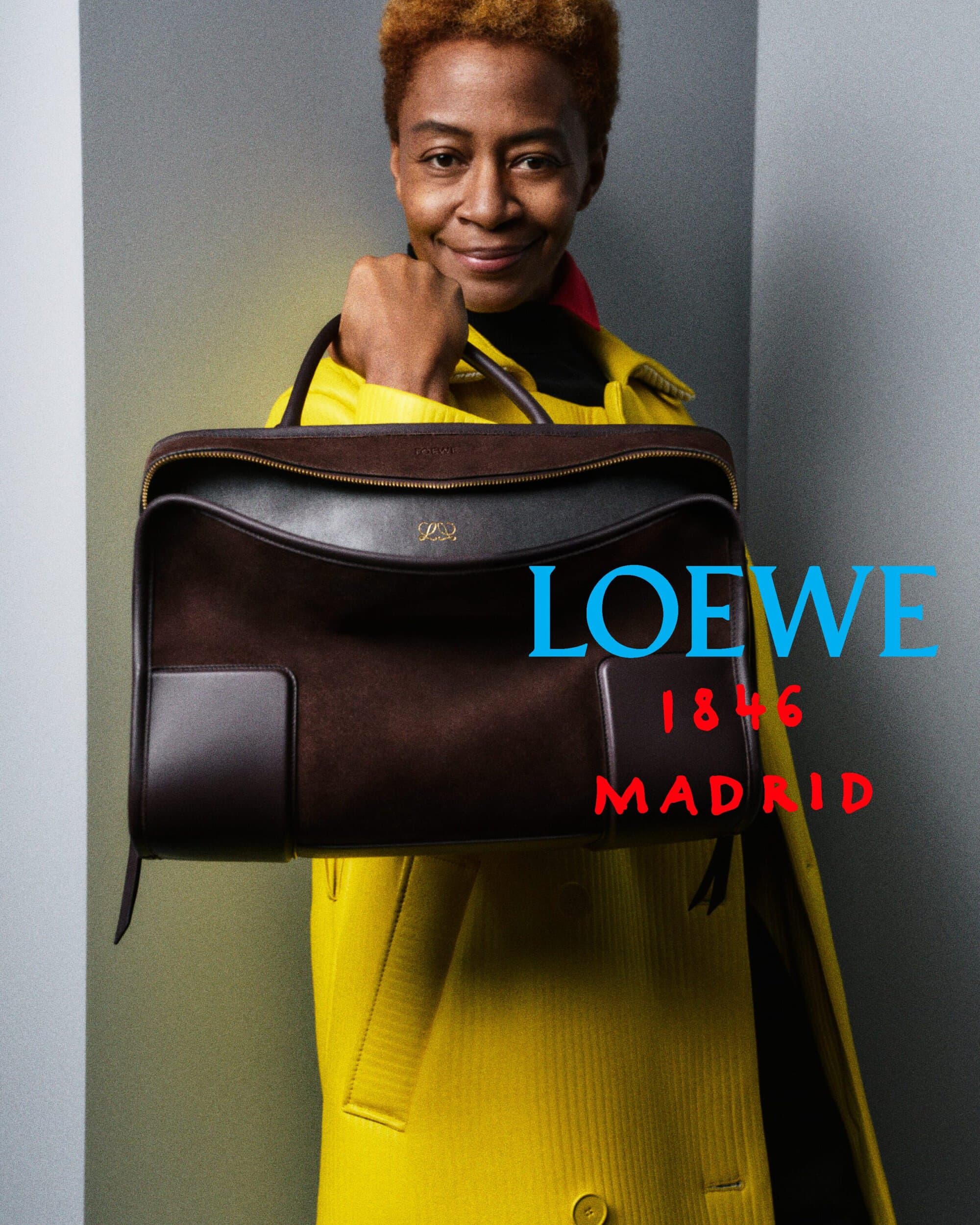 Kara Walker wearing a bright yellow coat and holding a large dark brown Loewe handbag against a studio backdrop.