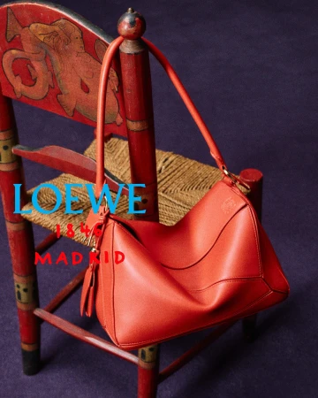 Orange leather shoulder bag draped over an ornate red painted wooden chair with a woven rush seat against a dark purple background.