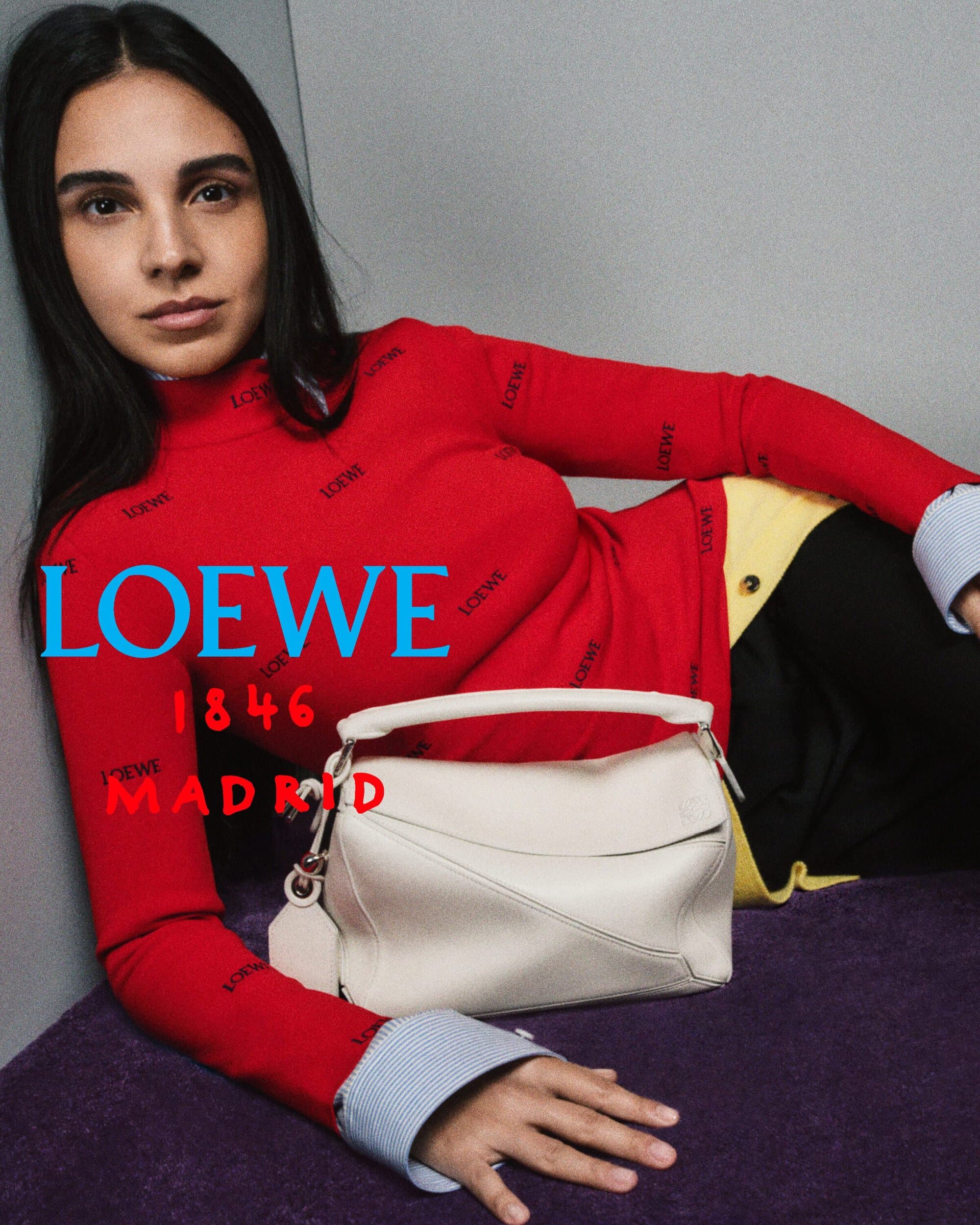 Salma Abu Deif reclining in a red turtleneck sweater beside an off-white leather handbag against a grey corner backdrop.