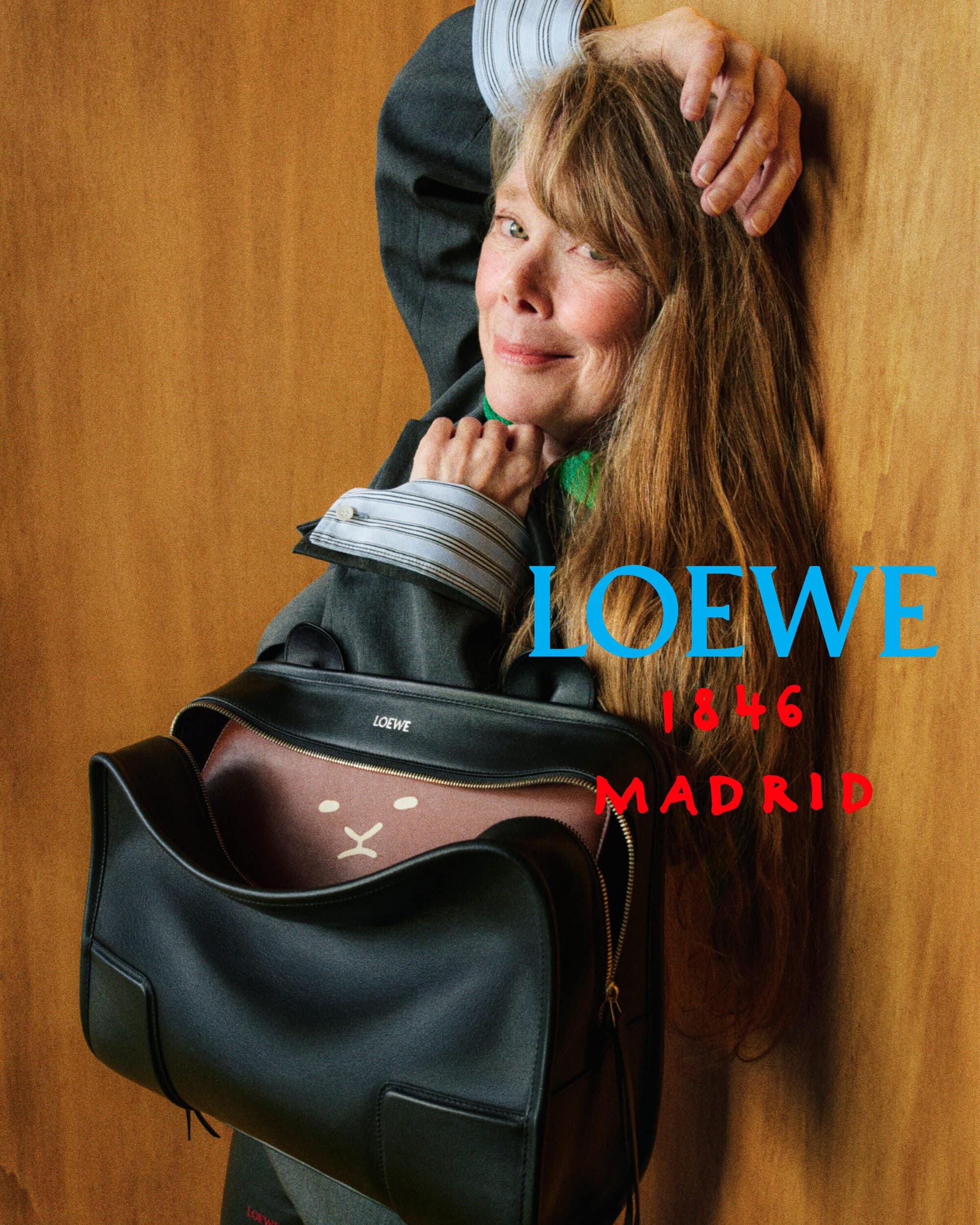 Actress Sissy Spacek rests against a wooden wall holding a large black leather Loewe handbag in a tailored grey jacket.