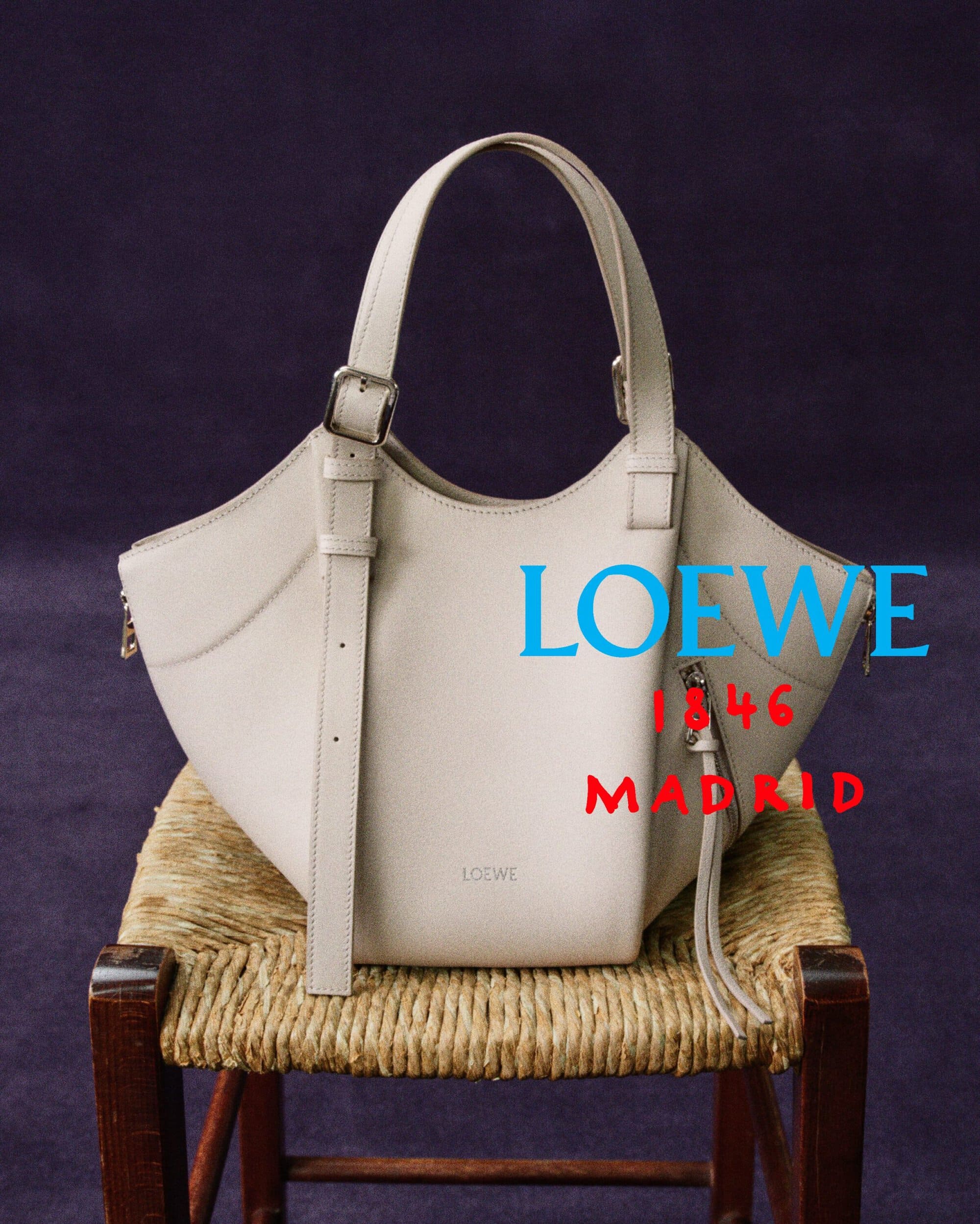 Off-white Loewe leather handbag placed on a rustic rush-seat chair against a dark purple backdrop.