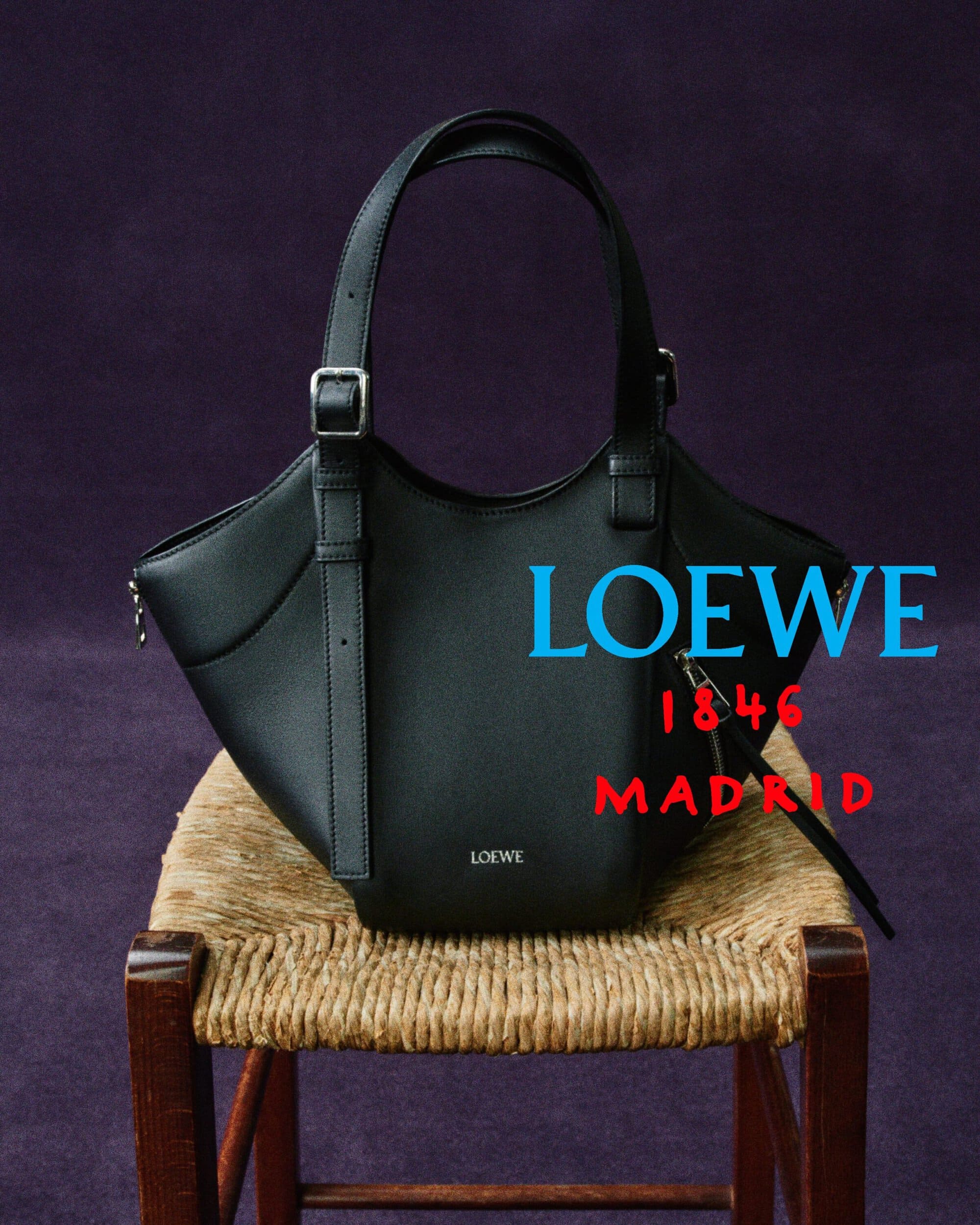 Black leather Loewe handbag resting on a rustic wooden chair with a woven rush seat against a deep purple studio backdrop.