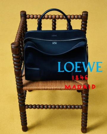 Dark navy suede handbag displayed on a turned wooden bobbin chair with a woven rush seat against a solid yellow backdrop.