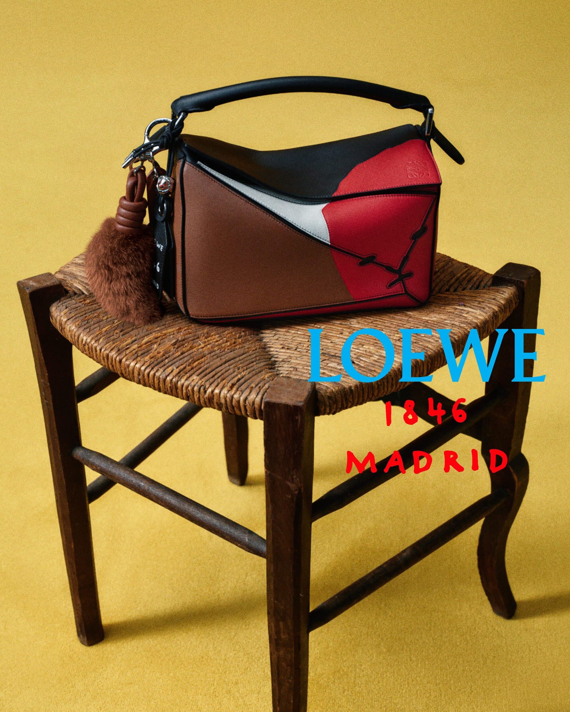 Loewe colorblocked leather handbag with a plush charm sitting on a rustic woven wooden chair against a yellow background.