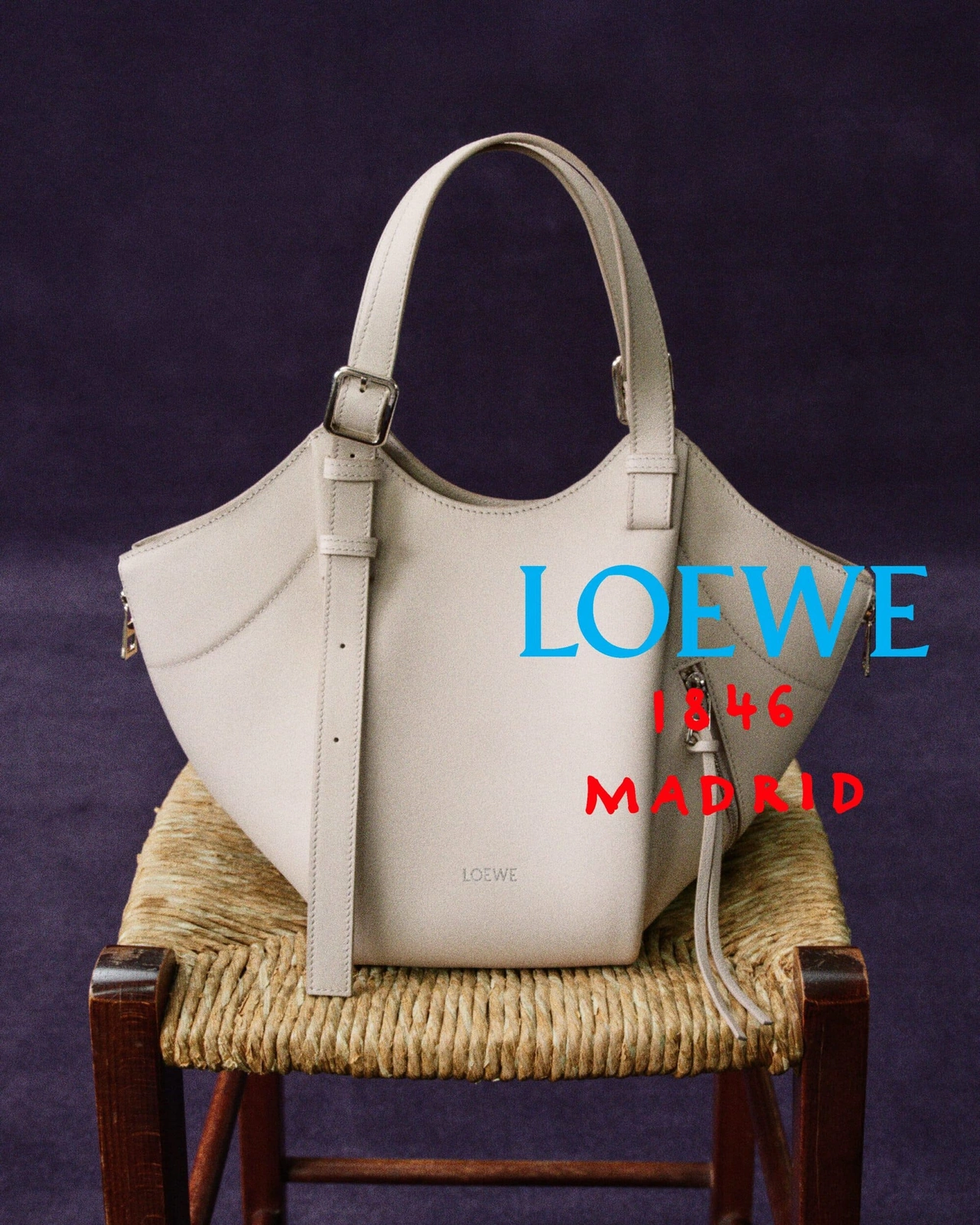Off-white Loewe leather handbag placed on a rustic rush-seat chair against a dark purple backdrop.