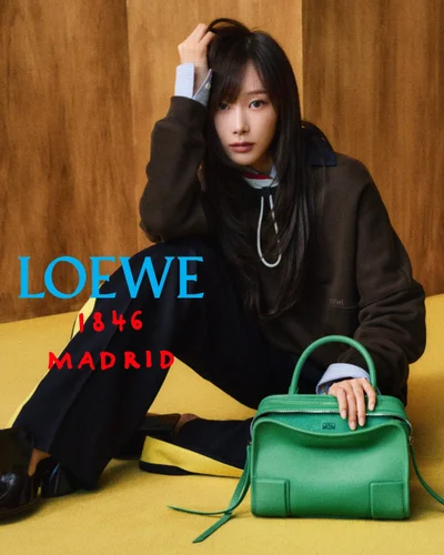 Giselle seated on a yellow floor in front of a wood wall, resting her hand on a vibrant green leather Loewe handbag.