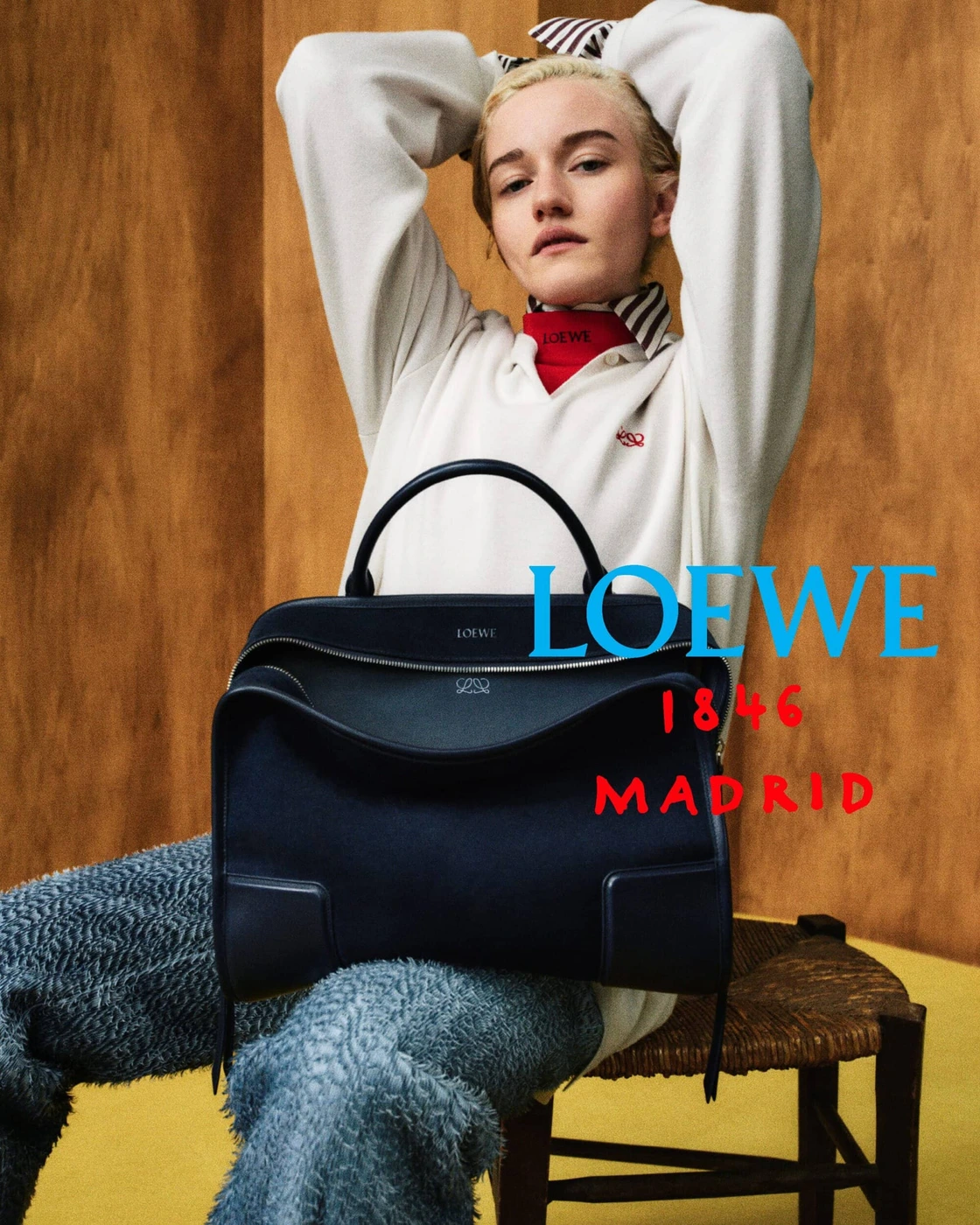Model seated on a wooden chair holding a large dark Loewe handbag against a wood-panel wall with campaign typography.