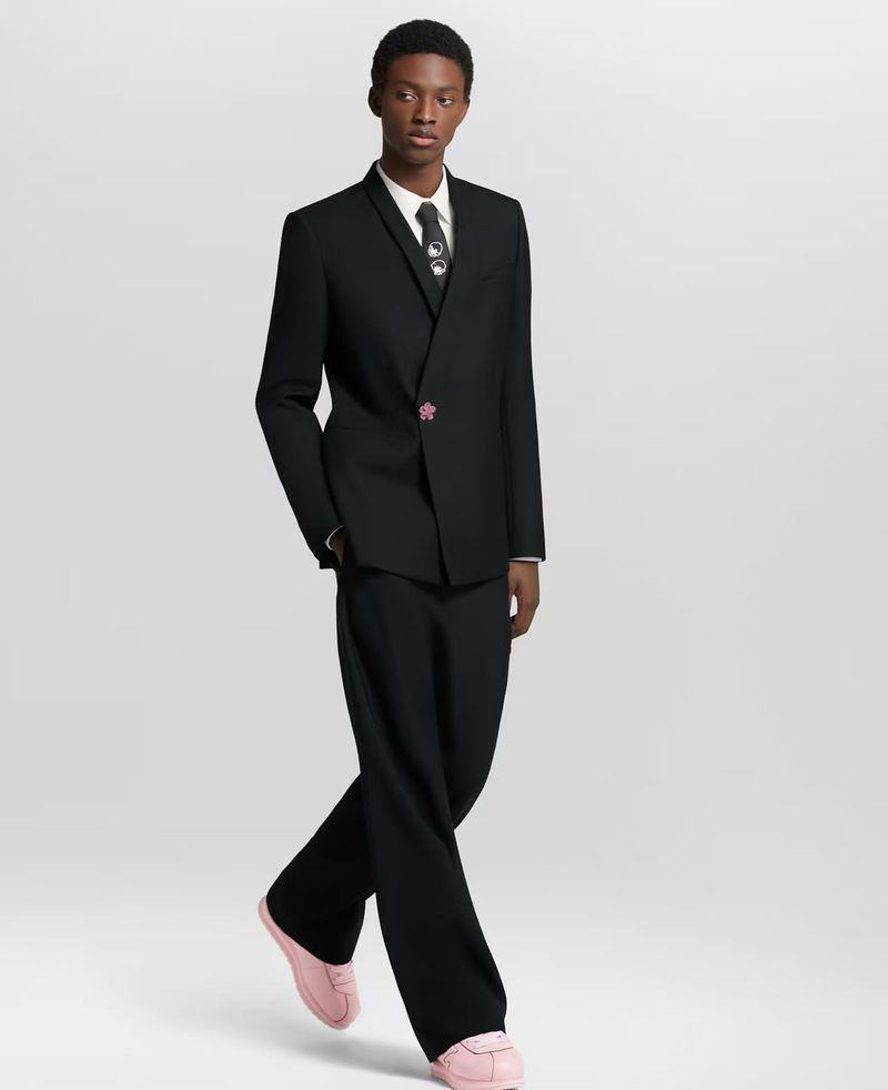 Young Black male fashion model poses in a black suit, white dress shirt, black tie, and pink sneakers against a light gray backdrop.