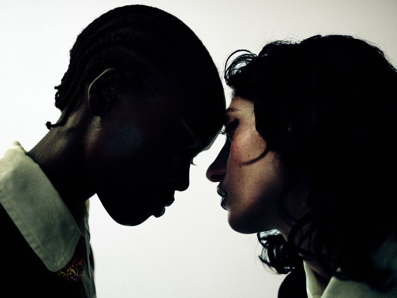 Close-up profile portrait of two women facing each other forehead to forehead against a pale illuminated background.