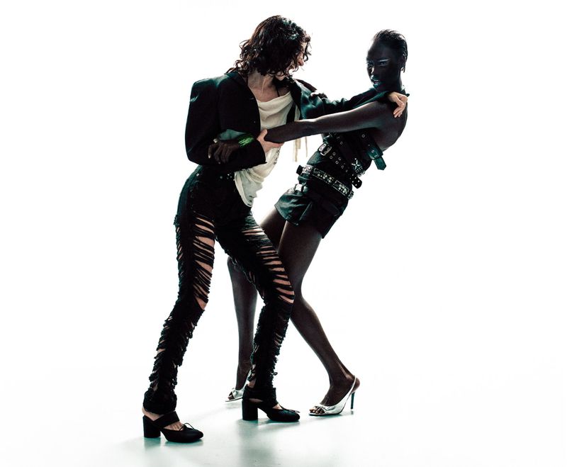 Two fashion models in distressed black garments and silver heels posing dynamically against a bright white background.